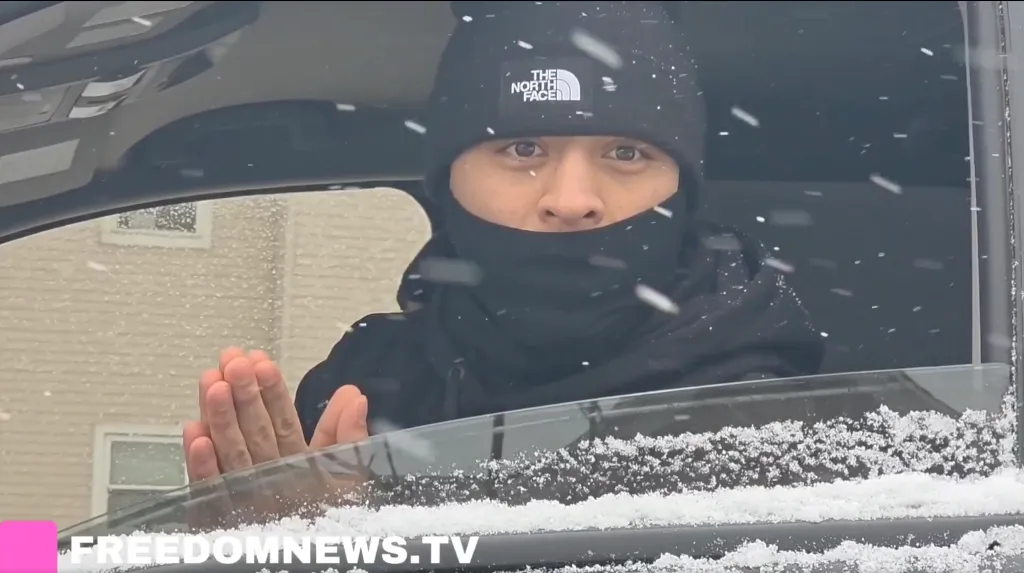 A person bundled in winter gear, wearing a black beanie and balaclava, looks out of a snowy car window with their hands pressed together as if in prayer.